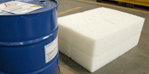A close-up of a TDI chemical drum with safety labels, next to a block of polyurethane foam.